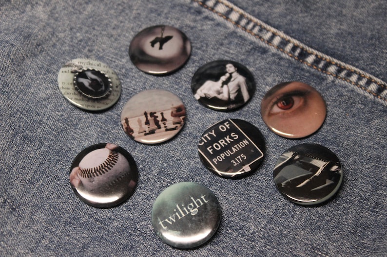 May include: Nine round buttons with various images related to the Twilight saga. The buttons feature images of a baseball, a chessboard, a sign that reads "City of Forks Population 3175", a silhouette of a person hanging from a tree, a close-up of an eye, a couple embracing, and the word "twilight" in a stylized font.