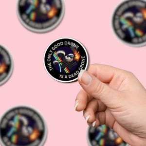 Pode incluir: Adesivo redondo com o texto "THE ONLY GOOD DRONE IS A DEAD DRONE". Apresenta um drone de desenho animado com óculos de sol, armas e um rastro de arco-íris. O fundo é azul escuro com uma explosão de fogo.