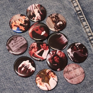 May include: A collection of round, black and white buttons featuring images of people and text. One button reads "Mystic Falls Virginia". Another button has the text "Stefan Salvatore". The buttons are arranged on a denim surface.