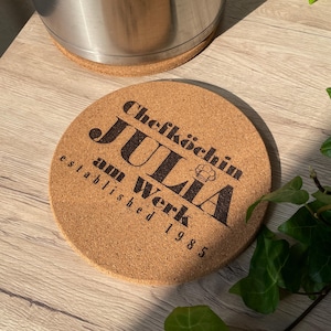 Könnte beinhalten: Ein runder Korkuntersetzer mit dem Text "ChefkÃ¶chin Julia am Werk established 1985" in schwarzer Schrift.