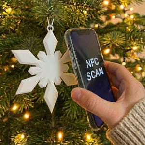Könnte beinhalten: Ein weißer Schneeflocken-Weihnachtsschmuck hängt an einem geschmückten Baum. Ein Smartphone mit der Aufschrift "NFC SCAN" wird in der Nähe des Schmucks gehalten. Der Hintergrund ist verschwommen mit Weihnachtslichtern.