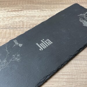Könnte beinhalten: Schwarzes Schiefer-Serviertablett mit einem weißen, geätzten Design eines Kolibris und eines blühenden Zweigs. Der Name "Julia" ist in der Mitte des Tabletts eingeätzt.
