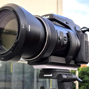 以下が含まれることがあります： 大きなズームレンズとレンズフードを装着した黒いNikon製デジタルブリッジカメラ。黒いCullmann製三脚ヘッドに固定されています。カメラ本体にはテクスチャ加工されたグリップ、側面のズームコントロールレバー、および側面にストラップが取り付けられています。