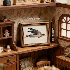 Puede incluir: Una impresión en miniatura enmarcada con una ilustración de un pez volador con alas negras y detalles rojos. La impresión mide 7,5 cm de ancho y 6,09 cm de alto, exhibida en una estantería de madera dentro de una casa de muñecas.