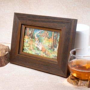 May include: A rectangular, dark brown wooden picture frame with a colorful illustration of a forest scene. The frame is angled, with a glass of amber liquid and a white cylinder in the background. The frame is resting on a wooden surface.