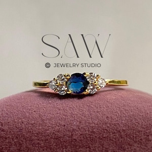 May include: A gold ring with a large, round, dark blue gemstone in the center, flanked by smaller, clear gemstones. The ring is set on a dark pink velvet surface. The text "SAW JEWELRY STUDIO" is visible in the background.