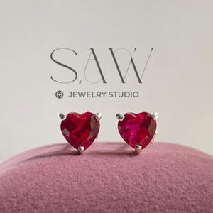 Peut inclure: Boucles d'oreilles en forme de cœur avec des rubis rouges et des montures argentées. Elles sont présentées sur un coussin en velours rose. Le texte "SAW JEWELRY STUDIO" est visible.