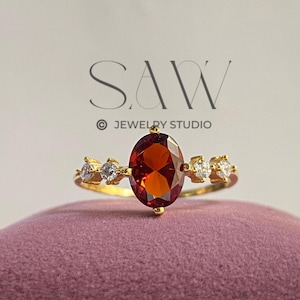 May include: A gold ring featuring an oval, reddish-brown gemstone as the centerpiece, flanked by smaller, clear stones. The ring is set on a pink velvet surface, with the text "SAW JEWELRY STUDIO" visible in the background.