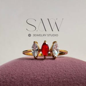 Könnte beinhalten: Ein goldfarbener Ring mit drei tropfenförmigen Edelsteinen. Der mittlere Stein ist rot, flankiert von zwei klaren, weißen Steinen. Der Ring liegt auf einer violetten Samtoberfläche. Im Hintergrund ist der Text "SAW JEWELRY STUDIO" zu sehen.