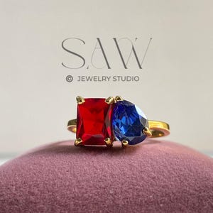 Może przedstawiać: Złoty pierścionek z czerwonym prostokątnym i niebieskim kamieniem w kształcie gruszki. Pierścionek jest prezentowany na różowej aksamitnej powierzchni. Tekst "SAW JEWELRY STUDIO" jest widoczny w tle.