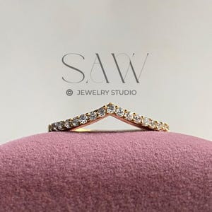 以下が含まれることがあります： 小さな透明な宝石がセットされた、金色のV字型のリング。リングはピンク色のベルベットの表面に展示されています。「SAW JEWELRY STUDIO」の文字が背景に見えます。
