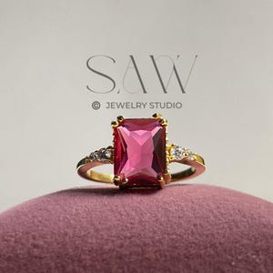 Puede incluir: Un anillo de oro con una gran piedra preciosa rosa rectangular. El anillo tiene pequeñas piedras claras a ambos lados de la piedra principal. Se exhibe sobre una superficie de terciopelo malva. El texto "SAW JEWELRY STUDIO" es visible.