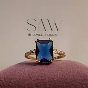 May include: A gold ring featuring a large, rectangular, deep blue gemstone. The ring has small, clear stones flanking the main stone. It is displayed on a maroon velvet surface. The text "SAW JEWELRY STUDIO" is visible.
