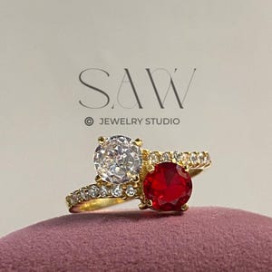 May include: A gold ring featuring two gemstones: a large, clear, round-cut stone and a smaller, red, round-cut stone. The ring is embellished with smaller, clear stones. The text "SAW JEWELRY STUDIO" is visible in the background.