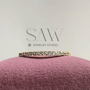 Pode incluir: Um delicado anel de ouro com uma fileira contínua de pequenas pedras preciosas transparentes. O anel repousa sobre uma superfície de veludo malva. O texto "SAW JEWELRY STUDIO" é visível.