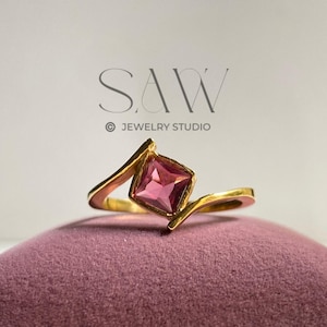 May include: A gold ring with a square, pink gemstone. The ring's band curves around the stone, creating a unique design. The ring is displayed on a pink velvet surface. The text "SAW JEWELRY STUDIO" is visible in the background.