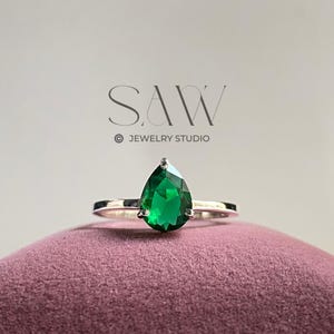 以下が含まれることがあります： シルバーリング、ペアシェイプのエメラルドグリーンの宝石付き。リングは濃いピンク色のベルベットの表面に置かれています。「SAW JEWELRY STUDIO」のロゴが背景に見えます。