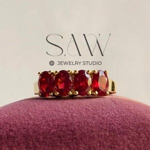 Puede incluir: Un anillo de oro con cinco piedras preciosas rojas ovaladas. El anillo se muestra sobre una superficie de terciopelo morado oscuro, con el logotipo del estudio de joyería en el fondo. Las piedras preciosas están espaciadas uniformemente.