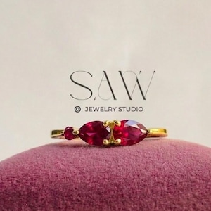 May include: A gold ring featuring three vibrant red gemstones. The ring has a delicate band and the gemstones are set in a row. The ring is displayed on a purple surface, with the text "SAW JEWELRY STUDIO" in the background.