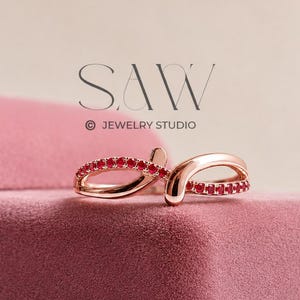 May include: A rose gold ring featuring a crossover design with small, round, vibrant red stones set along the band. The ring is positioned on a dusty pink velvet jewelry box. The text SAW JEWELRY STUDIO is visible above the ring.