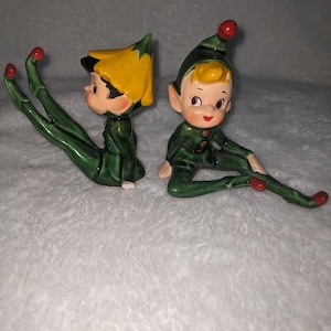Peut inclure: Deux figurines d'elfes en céramique verte et jaune avec des chapeaux et des chaussures rouges. Les elfes sont assis, les jambes tendues.