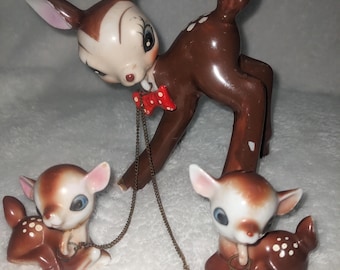May include: A set of three ceramic deer figurines. The mother deer is brown with white spots and wears a red bow tie. The two fawn deer are brown with white spots and are connected to the mother deer by a gold chain.