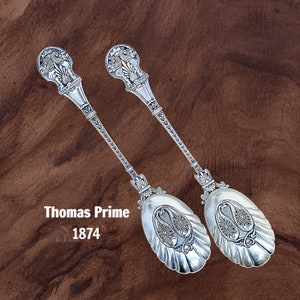 May include: Two silver spoons with ornate handles and floral designs on the bowl. The handles feature a bird design. The spoons are on a brown wooden surface. The text "Thomas Prime 1874" is visible below the spoons.