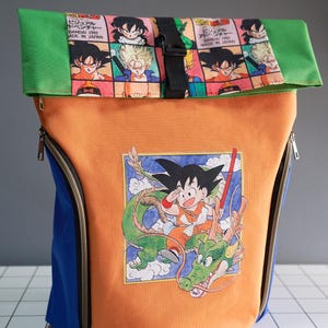 Peut inclure: Sac à dos orange avec des panneaux bleus et verts. L'avant présente une illustration colorée d'un personnage d'anime chevauchant un dragon. Le rabat supérieur est vert, avec un intérieur à motifs avec des images de personnages d'anime. Il a une boucle noire et deux fermetures éclair.