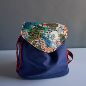 Op de afbeelding: Een marineblauwe rugzak met trekkoord en een klep met bloemenprint. De klep is gemaakt van een kleurrijke stof met een blauwe, groene, rode en witte bloemenprint.