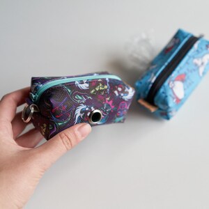 Könnte beinhalten: Zwei kleine, farbige, Reißverschlussbeutel mit einem Schlüsselring. Ein Beutel ist schwarz mit einem bunten, abstrakten Design. Der andere Beutel ist blau mit einem weißen und roten Cartoon-Design.