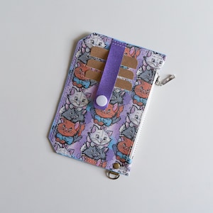 Puede incluir: Una cartera de color morado y blanco con cierre de cremallera y un patrón repetido de gatos de dibujos animados. La cartera tiene una correa morada con un botón blanco.