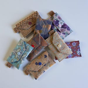 Può includere: Una collezione di otto piccoli portafogli colorati realizzati in sughero e tessuto. I portafogli hanno diversi motivi e design, tra cui motivi floreali, geometrici e astratti. I portafogli sono tutti chiusi e hanno una chiusura a bottone.