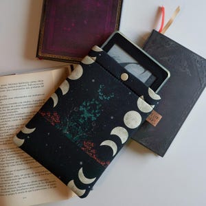 Op de afbeelding: Zwarte e-reader hoes met een maan- en sterrenpatroon. De hoes heeft een flap die met een knoop sluit. De hoes is gemaakt van een zachte, duurzame stof.