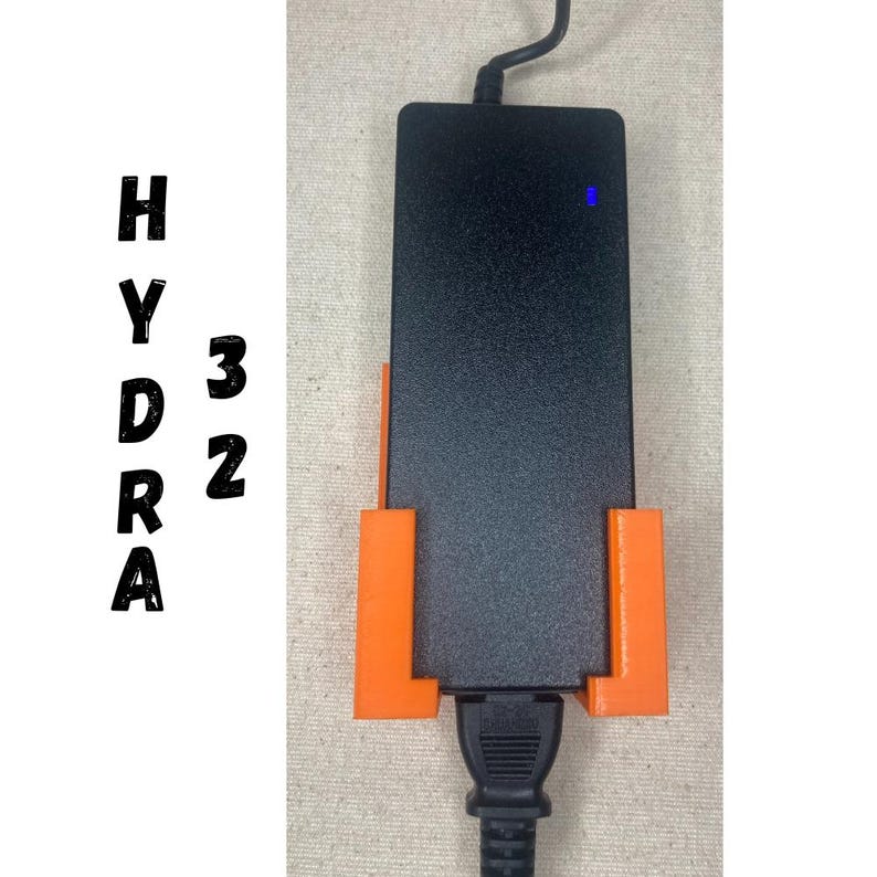 Puede incluir: Adaptador de corriente rectangular negro con un soporte naranja. El adaptador tiene una luz azul y un cable de alimentaci&oacute;n negro. El texto "HYDRA 32" est&aacute; impreso verticalmente en el lado izquierdo de la imagen.