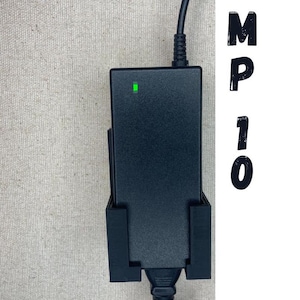 Op de afbeelding: Een zwarte stroomadapter met een groene indicatorlamp, gemonteerd aan een muur met een wit snoer dat is aangesloten. De tekst "MP10" is zichtbaar op de achtergrond.