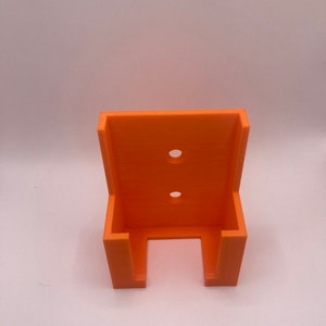 Puede incluir: Un soporte de pared naranja impreso en 3D con dos agujeros circulares y dos soportes laterales. El soporte est&aacute; dise&ntilde;ado para sujetar un objeto y est&aacute; hecho de pl&aacute;stico naranja brillante.