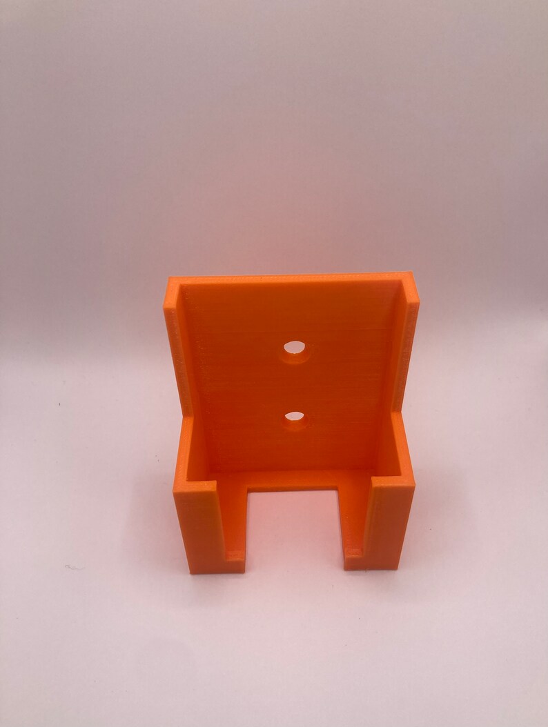 Puede incluir: Un soporte de pared naranja impreso en 3D con dos agujeros circulares y dos soportes laterales. El soporte est&aacute; dise&ntilde;ado para sujetar un objeto y est&aacute; hecho de pl&aacute;stico naranja brillante.
