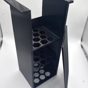 Könnte beinhalten: Eine schwarze, 3D-gedruckte Aufbewahrungsbox mit einer Scharniertür. Der Innenraum verfügt über mehrere Regale mit sechseckigen Aussparungen. Die Box ist für die Aufnahme kleiner Gegenstände konzipiert. Die Box besteht aus schwarzem Kunststoff.