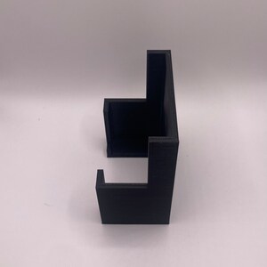 Puede incluir: Un objeto negro impreso en 3D con un dise&ntilde;o angular &uacute;nico. El objeto tiene una base rectangular con dos secciones elevadas perpendiculares, creando un soporte. El material parece ser pl&aacute;stico mate.