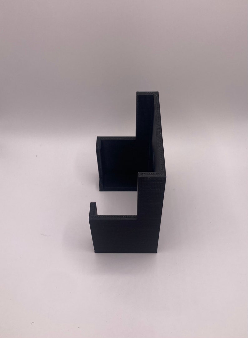 Puede incluir: Un objeto negro impreso en 3D con un dise&ntilde;o angular &uacute;nico. El objeto tiene una base rectangular con dos secciones elevadas perpendiculares, creando un soporte. El material parece ser pl&aacute;stico mate.