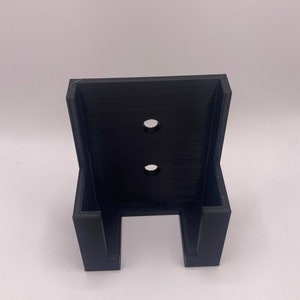 Puede incluir: Un soporte de pared negro impreso en 3D con dos agujeros circulares y una abertura en forma de U en la parte inferior. El soporte est&aacute; dise&ntilde;ado para sujetar un art&iacute;culo y est&aacute; hecho de un material negro mate.