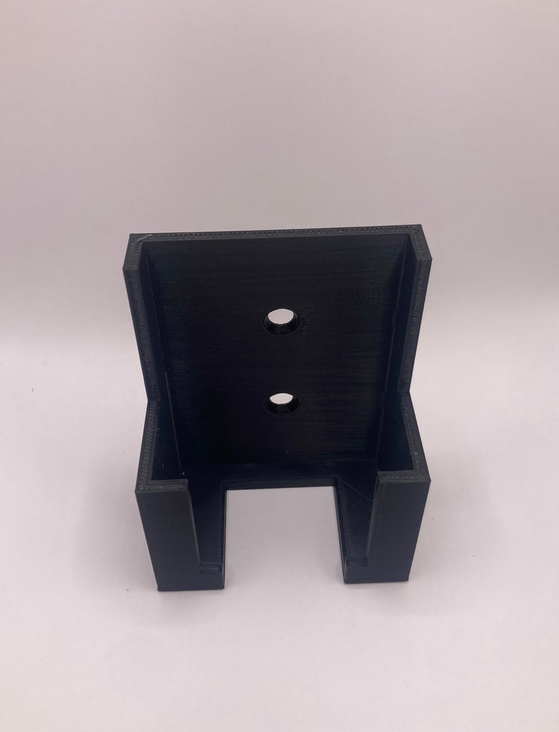 Puede incluir: Un soporte de pared negro impreso en 3D con dos agujeros circulares y una abertura en forma de U en la parte inferior. El soporte est&aacute; dise&ntilde;ado para sujetar un art&iacute;culo y est&aacute; hecho de un material negro mate.