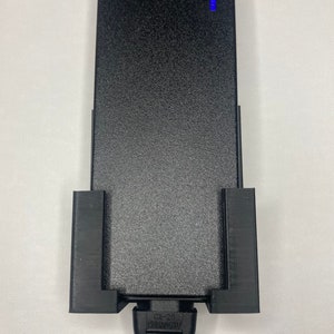 Puede incluir: Un adaptador de corriente negro con una superficie texturizada y una luz indicadora azul. El adaptador se sujeta con un soporte negro. El cable de alimentaci&oacute;n es negro y tiene un enchufe de dos clavijas.