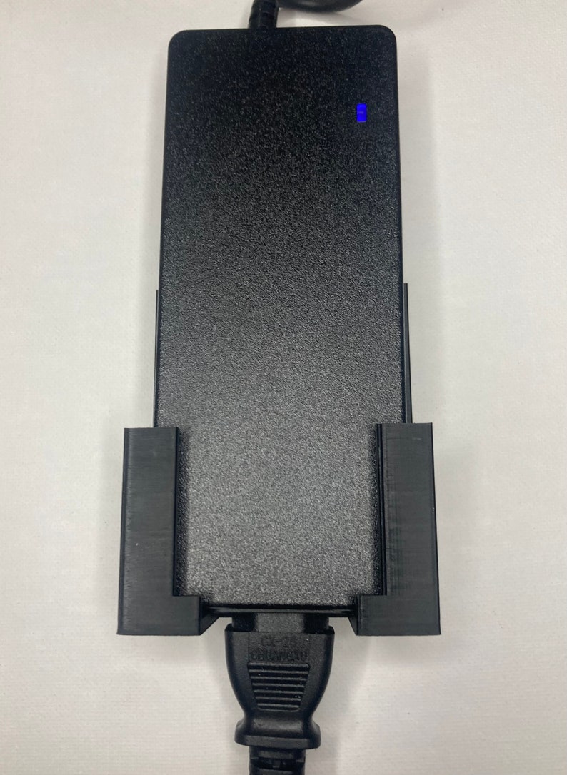 Puede incluir: Un adaptador de corriente negro con una superficie texturizada y una luz indicadora azul. El adaptador se sujeta con un soporte negro. El cable de alimentaci&oacute;n es negro y tiene un enchufe de dos clavijas.