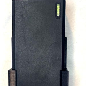 May include: A rectangular black power adapter brick secured within a black plastic mounting bracket. The adapter features a small green LED indicator light near the top and a series of six small circular indentations arranged in a triangular pattern.