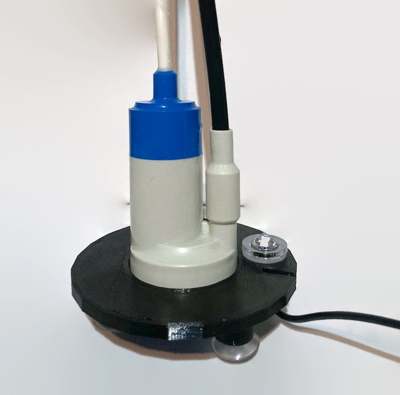 May include: A small, white submersible pump with a blue top and a black wire. The pump is mounted on a black plastic base with a clear plastic cap on the side.