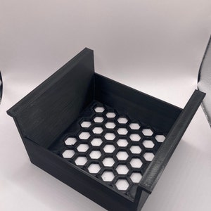 Puede incluir: Una caja de almacenamiento de plástico negro con una rejilla en forma de panal en la parte inferior. La caja tiene un borde elevado en un lado.