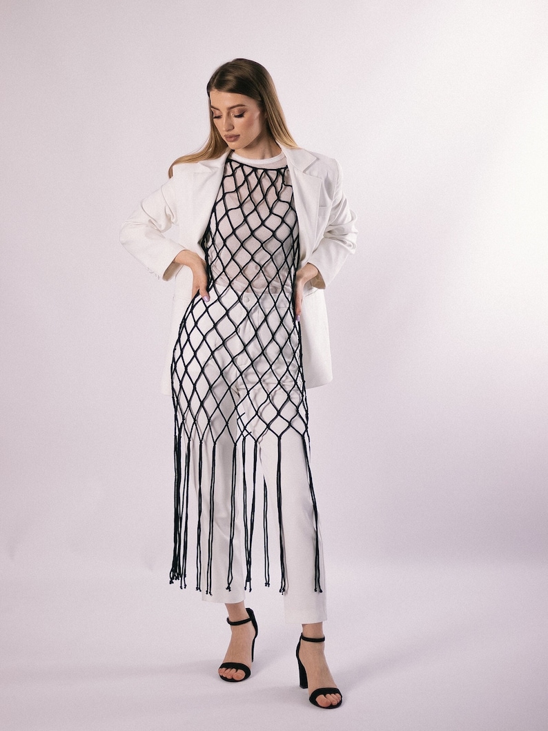 May include: A woman wearing a white blazer, white pants, and a black mesh top with fringe. She is standing on a white background.