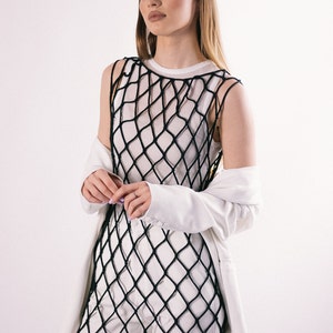 May include: A woman wearing a white top with a black fishnet overlay and a white blazer. She is standing in front of a white background.