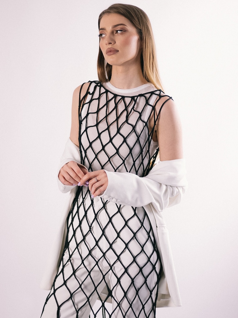 May include: A woman wearing a white top with a black fishnet overlay and a white blazer. She is standing in front of a white background.